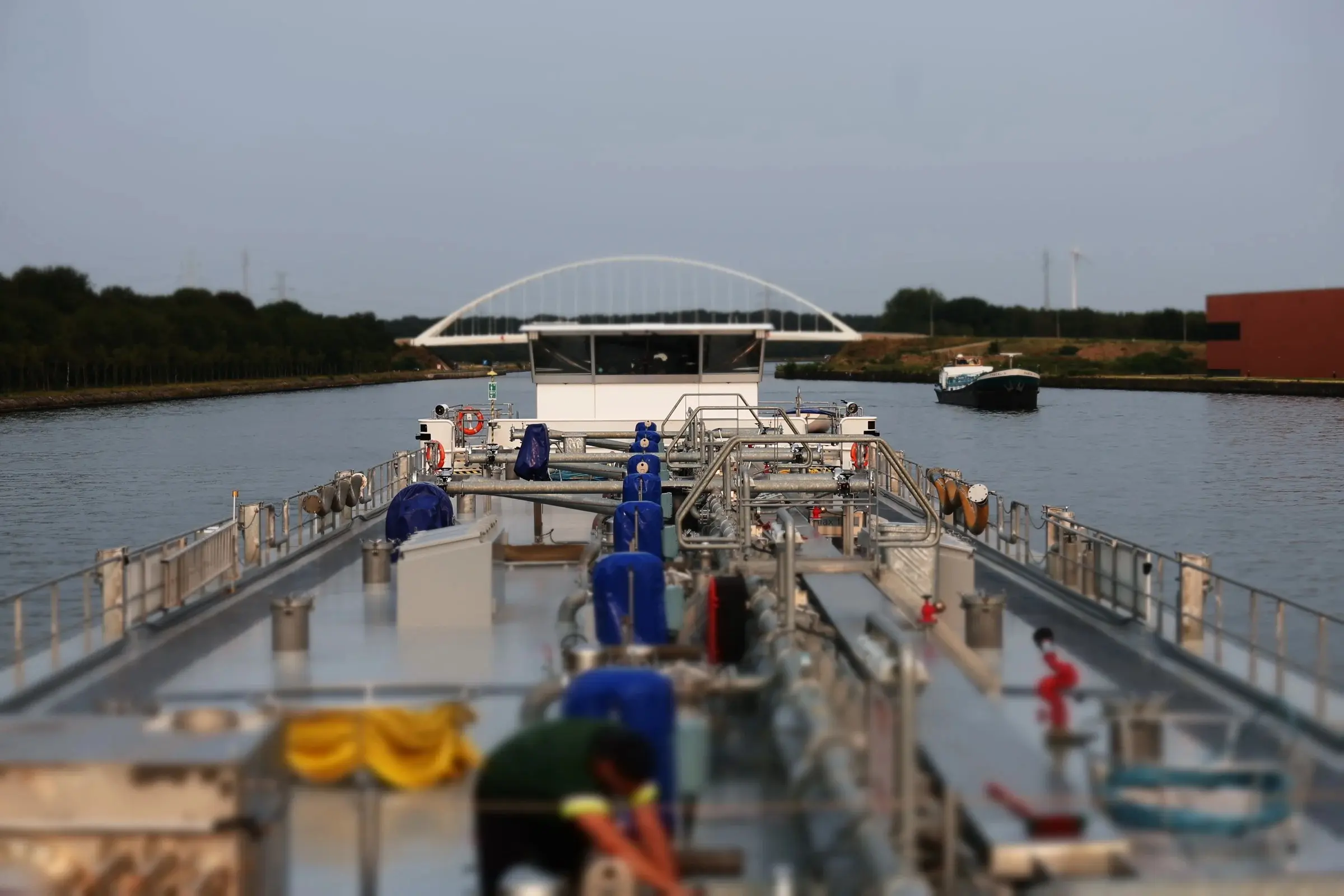 A tanker’s deck and wheelhouse on a canal, with an arched bridge behind.